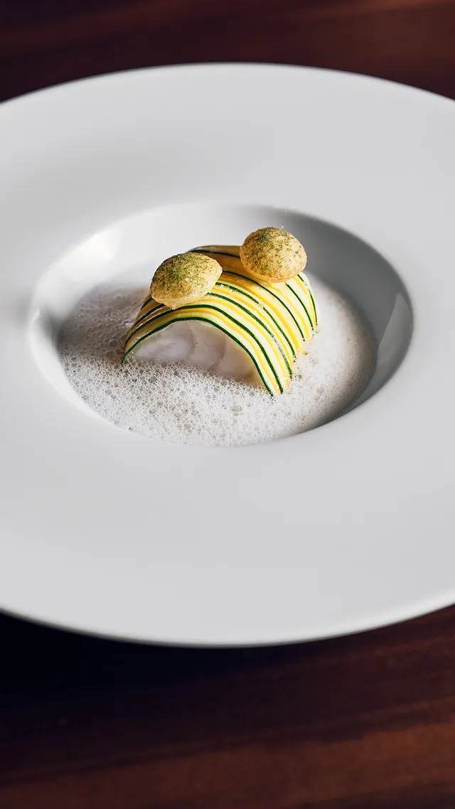 Gourmetgericht mit Schaum, Gemüsebändern und Garnierungen auf weißem Teller auf Holztisch
